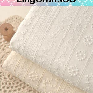 May include: Two folded pieces of white and off-white cotton fabric featuring vertical stripes of intricate floral embroidery and eyelet patterns. The fabric displays a textured surface with repeating botanical motifs and delicate needlework details.