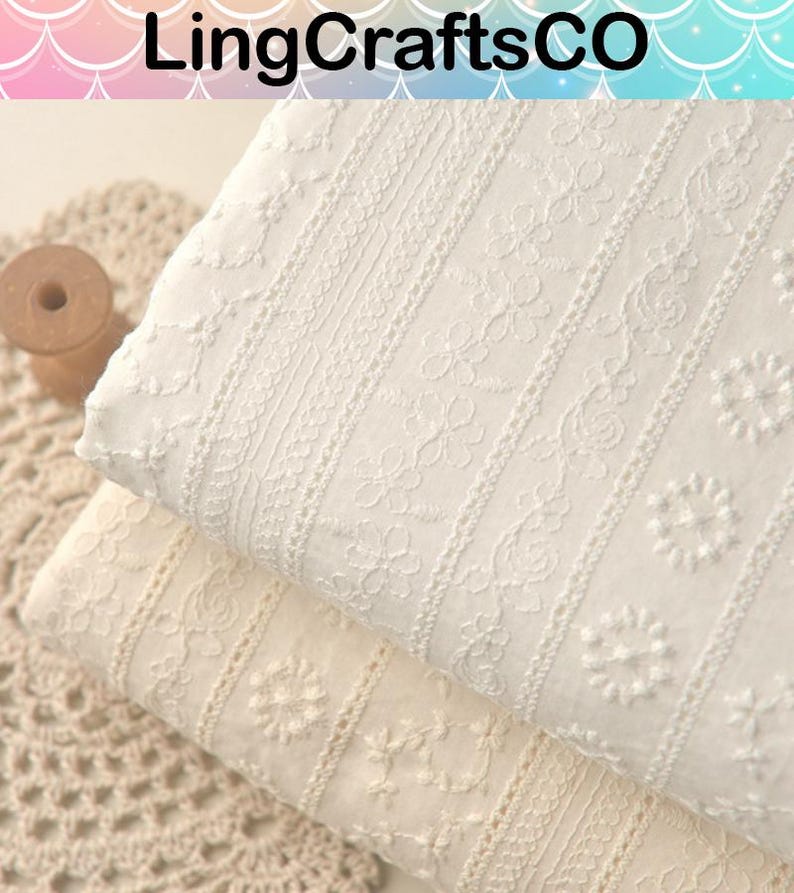 May include: Two folded pieces of white and off-white cotton fabric featuring vertical stripes of intricate floral embroidery and eyelet patterns. The fabric displays a textured surface with repeating botanical motifs and delicate needlework details.