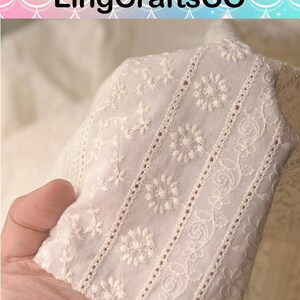 May include: Off-white fabric featuring intricate white floral embroidery and vertical eyelet lace detailing. The textile displays a repeating pattern of small blossoms, swirling vines, and circular motifs arranged in distinct, parallel decorative bands.