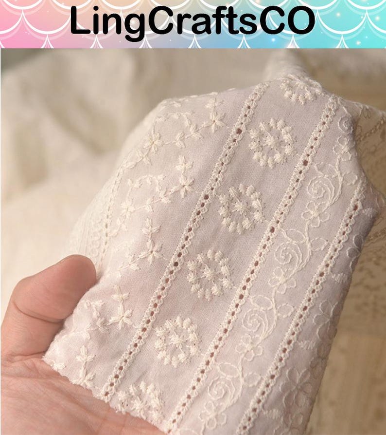May include: Off-white fabric featuring intricate white floral embroidery and vertical eyelet lace detailing. The textile displays a repeating pattern of small blossoms, swirling vines, and circular motifs arranged in distinct, parallel decorative bands.