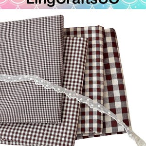 May include: Four folded fabric squares featuring various sizes of brown and white gingham check patterns, layered together. A thin, white, scalloped lace trim with small, evenly spaced white dots is draped diagonally across the fabric stack.