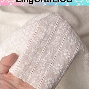 May include: A white fabric featuring intricate eyelet embroidery with repeating floral patterns and vertical decorative bands. The textile displays a delicate, semi-sheer texture with detailed needlework throughout the surface, labeled LingCraftsCO.