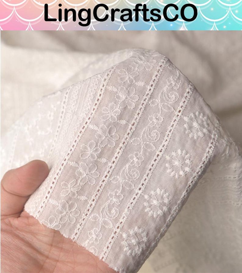 May include: A white fabric featuring intricate eyelet embroidery with repeating floral patterns and vertical decorative bands. The textile displays a delicate, semi-sheer texture with detailed needlework throughout the surface, labeled LingCraftsCO.