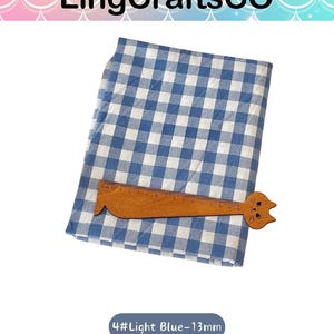 Puede incluir: Una pieza de tela doblada con un patr&oacute;n de cuadros vichy en azul y blanco. Una regla de madera con forma de gato est&aacute; colocada sobre la tela, mostrando una escala de medici&oacute;n de hasta 25 cent&iacute;metros. El texto 4#Light Blue-13mm es visible.