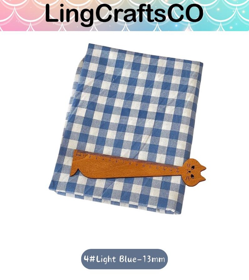 Puede incluir: Una pieza de tela doblada con un patr&oacute;n de cuadros vichy en azul y blanco. Una regla de madera con forma de gato est&aacute; colocada sobre la tela, mostrando una escala de medici&oacute;n de hasta 25 cent&iacute;metros. El texto 4#Light Blue-13mm es visible.