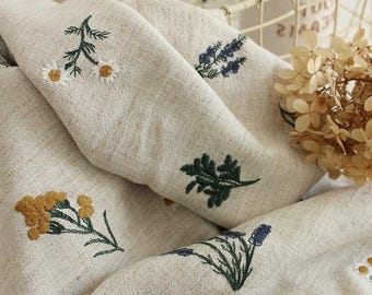 May include: A beige fabric featuring embroidered botanical motifs including white daisies with yellow centers, dark green leafy sprigs, golden yellow wildflower clusters, and purple lavender stalks. The textile has a visible weave texture.