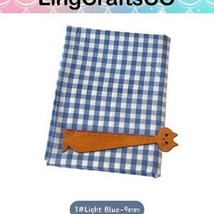 Puede incluir: Una tela doblada con un patr&oacute;n de cuadros vichy en azul y blanco. Una regla de madera con forma de gato est&aacute; colocada sobre la tela, mostrando una escala de medici&oacute;n de hasta 15 cm. El texto 3#Light Blue-9mm es visible debajo.