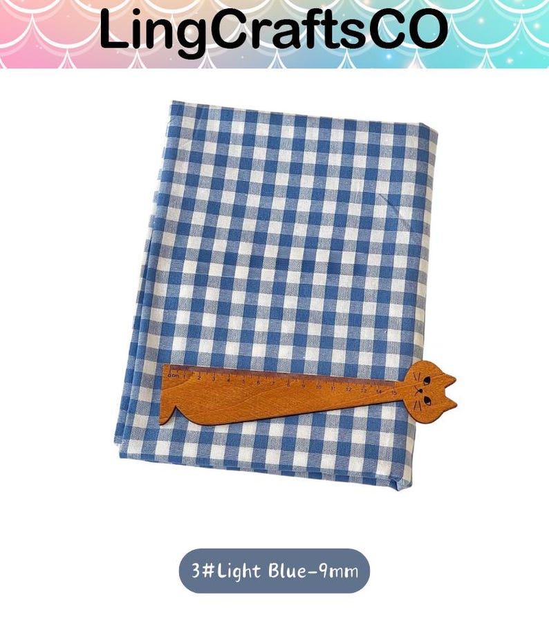 Puede incluir: Una tela doblada con un patr&oacute;n de cuadros vichy en azul y blanco. Una regla de madera con forma de gato est&aacute; colocada sobre la tela, mostrando una escala de medici&oacute;n de hasta 15 cm. El texto 3#Light Blue-9mm es visible debajo.