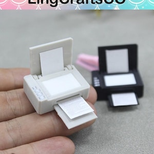 May include: Two miniature toy printers, one white and one black, with paper trays and a sheet of paper coming out of the white printer.