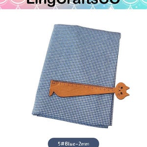 Puede incluir: Una pieza doblada de tela de cuadros vichy azul y blanca con una peque&ntilde;a regla de madera con forma de gato encima. La regla tiene marcas en cent&iacute;metros, mostrando una longitud de 15 cm. El texto 5#Blue-2mm es visible debajo de la tela.