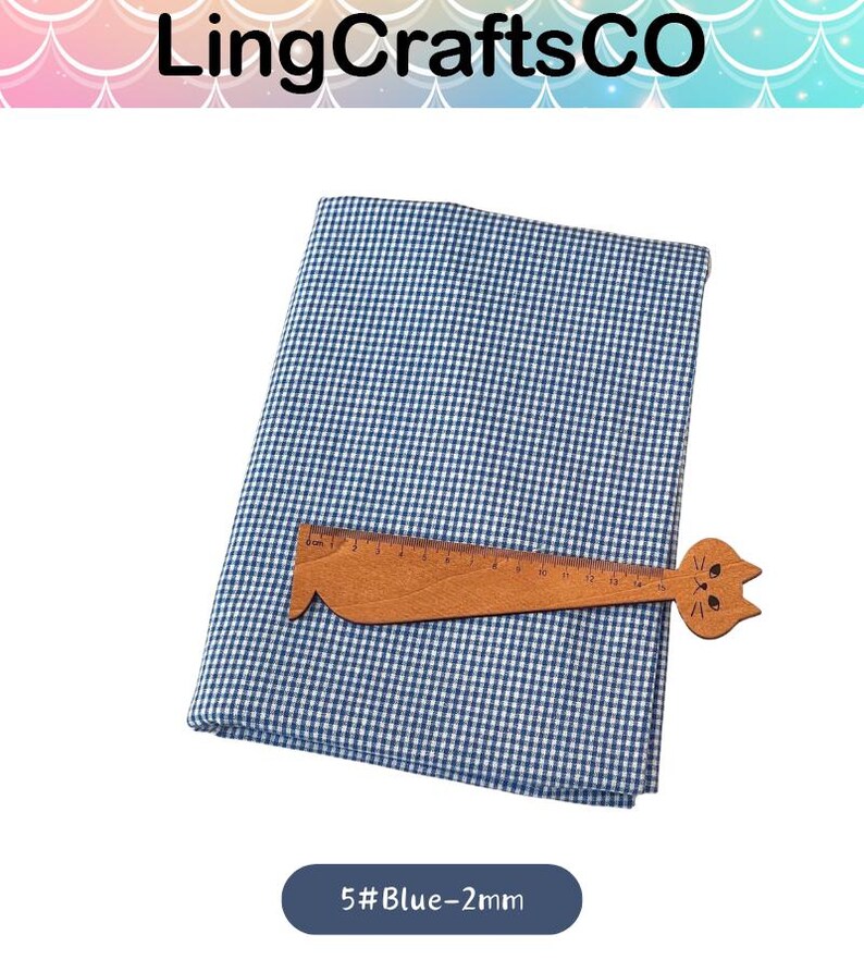 Puede incluir: Una pieza doblada de tela de cuadros vichy azul y blanca con una peque&ntilde;a regla de madera con forma de gato encima. La regla tiene marcas en cent&iacute;metros, mostrando una longitud de 15 cm. El texto 5#Blue-2mm es visible debajo de la tela.
