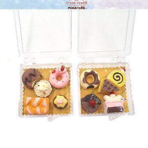 May include: A clear plastic box with a yellow and white patterned insert containing six miniature plastic pastries. The pastries include a donut, a cake, a roll, a croissant, and two other pastries. The box is labeled "Stone Tower Miniature".