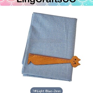 Puede incluir: Una pieza doblada de tela de cuadros vichy en azul claro y blanco. Una regla de madera con forma de gato est&aacute; colocada sobre la tela, mostrando una escala de medici&oacute;n de 0 a 16 cent&iacute;metros. El texto 1#Light Blue-2mm es visible debajo.