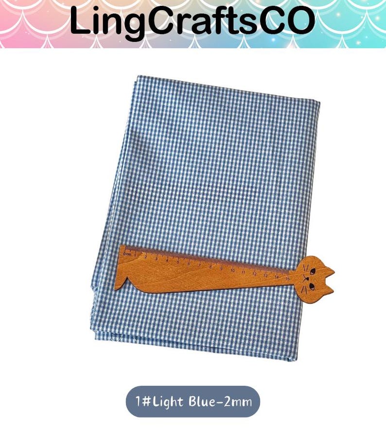 Puede incluir: Una pieza doblada de tela de cuadros vichy en azul claro y blanco. Una regla de madera con forma de gato est&aacute; colocada sobre la tela, mostrando una escala de medici&oacute;n de 0 a 16 cent&iacute;metros. El texto 1#Light Blue-2mm es visible debajo.