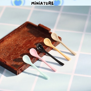 May include: Six miniature spoons in various colors, including light blue, pink, black, and yellow, are arranged on a wooden tray. The spoons are made of plastic and have a rounded shape. The spoons are part of a miniature kitchen set.