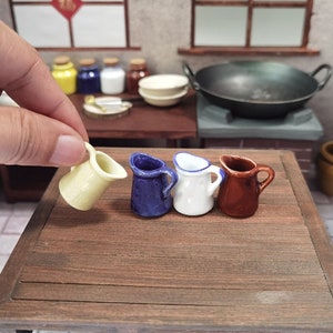 May include: A hand holds a small, yellow ceramic pitcher next to three other miniature pitchers. The other pitchers are blue, white, and brown. The pitchers have small handles and spouts, and are arranged on a wooden surface.