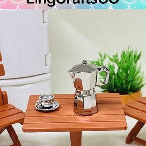 Może przedstawiać: Miniaturowa srebrna kawiarka i mała biała filiżanka z talerzykiem na brązowym drewnianym stole. Stół otoczony jest dwoma małymi drewnianymi krzesłami.