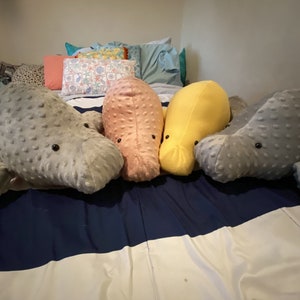 May include: Four plush manatee toys in different colors: gray, pink, yellow, and gray. The toys are lying on a striped bedspread.