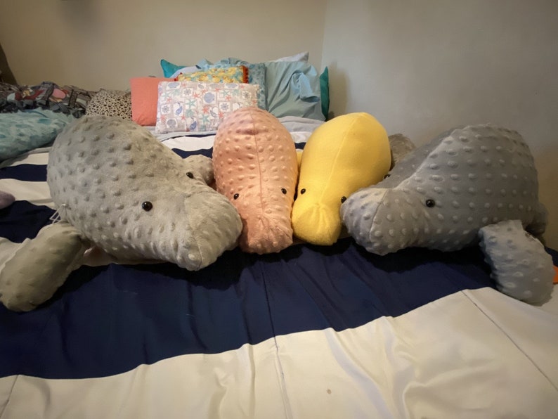 May include: Four plush manatee toys in different colors: gray, pink, yellow, and gray. The toys are lying on a striped bedspread.