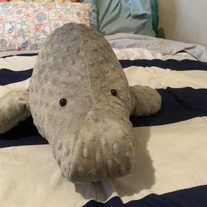 May include: A gray plush manatee toy with black eyes, lying on a white and blue striped bedspread.