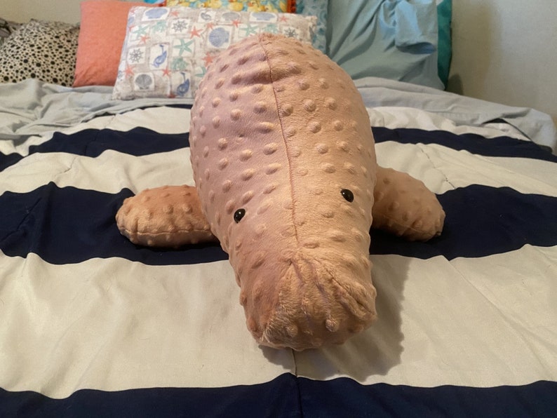 May include: A pink plush manatee toy with black eyes. The toy is lying on a striped blue and white blanket.