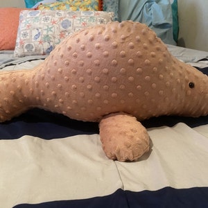 May include: A plush pink manatee shaped pillow with a textured surface. The pillow is lying on a striped bedspread.