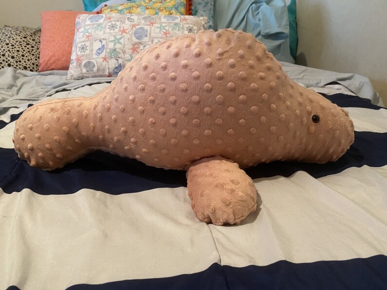 May include: A plush pink manatee shaped pillow with a textured surface. The pillow is lying on a striped bedspread.