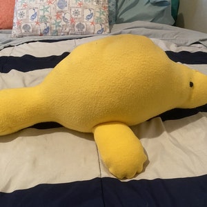 May include: A large yellow plush manatee toy lying on a striped bedspread. The manatee has a small black eye and a slightly upturned snout.