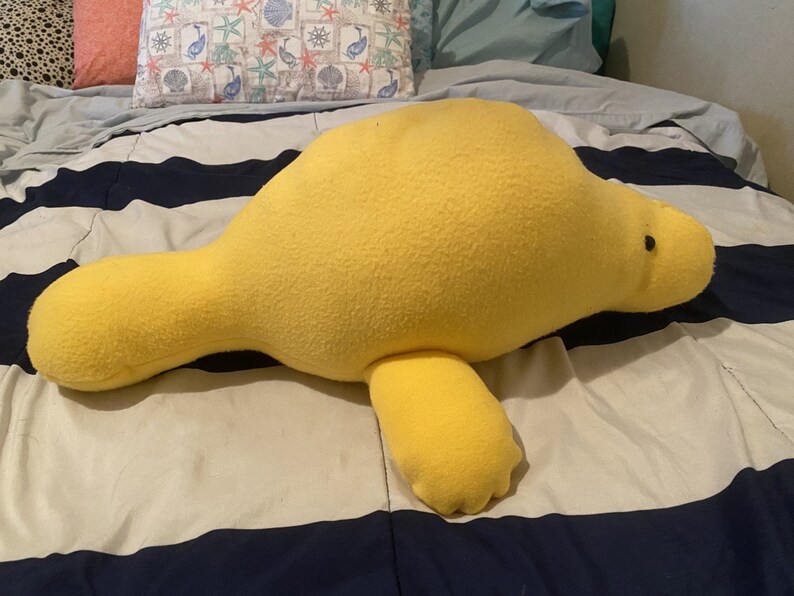 May include: A large yellow plush manatee toy lying on a striped bedspread. The manatee has a small black eye and a slightly upturned snout.