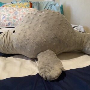 May include: A gray plush manatee toy with a textured surface. The manatee is lying on a striped bedspread with white and navy blue stripes.