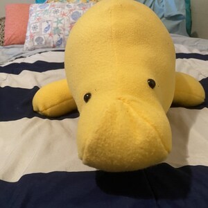 May include: A yellow plush manatee with black eyes. The manatee is lying on a white and blue striped bedspread.
