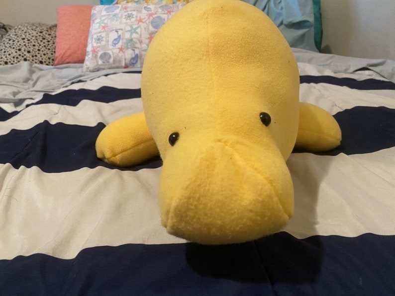 May include: A yellow plush manatee with black eyes. The manatee is lying on a white and blue striped bedspread.