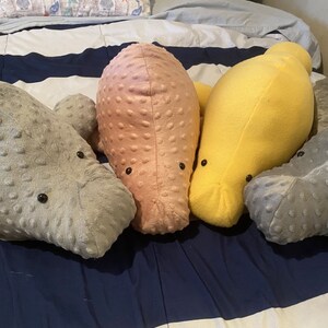 May include: Four plush manatee toys in different colors: gray, pink, yellow, and gray. The manatees are lying on a striped bedspread.