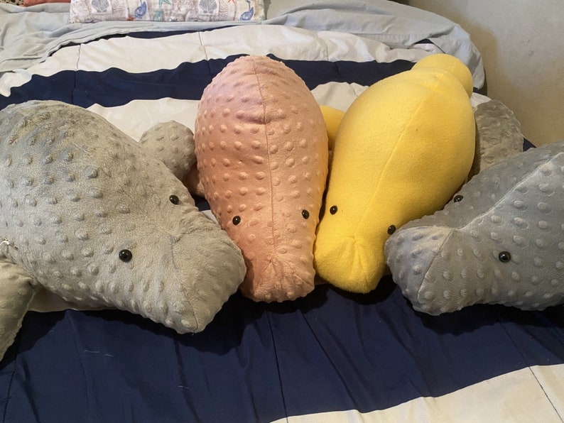 May include: Four plush manatee toys in different colors: gray, pink, yellow, and gray. The manatees are lying on a striped bedspread.