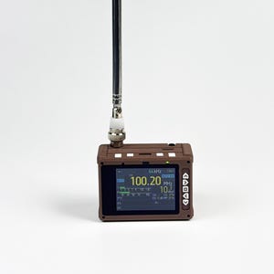 Puede incluir: Un receptor de radio portátil marrón con una antena telescópica negra larga y delgada que se extiende verticalmente desde la parte superior. El dispositivo cuenta con una pantalla digital rectangular que muestra la frecuencia 100.20 MHz y ajustes.