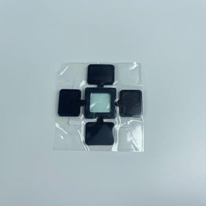 May include: A black, cross-shaped product with a central square and four rectangular extensions, packaged in clear plastic. The center square has a light blue tint. The product is set against a plain white background.