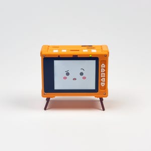 May include: A small orange electronic device with a rectangular screen displaying a cartoon face with sad eyes and pink cheeks. The device features a vertical row of five white navigation buttons on the right side and stands on two thin dark legs.