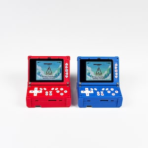 Puede incluir: Una consola de juegos portátil roja y otra azul, cada una con una pantalla que muestra el texto Attaky handheld gaming console, una cruceta direccional y varios botones de control incluyendo A, B, X e Y, con un puerto USB-C en el borde inferior.