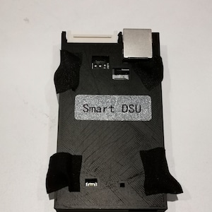 May include: A black Smart DSU device with a silver component on the top right corner. The device has a label that says "Smart DSU".