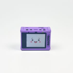 May include: A rectangular purple electronic device with a screen displaying a cartoon face with black eyes and a smiling mouth. The device features a black border around the screen, a small green indicator light, and black navigation buttons.