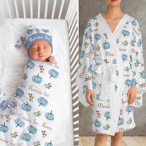 Può includere: Un bambino avvolto in una coperta bianca e con un cappello abbinato, entrambi decorati con zucche blu, foglie verdi e il nome "Thomas Lee". È presente anche un accappatoio bianco con un motivo di zucche e foglie abbinato e la parola "Mama".
