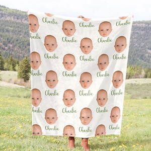 May include: A white blanket with a repeating pattern of a baby's face and the name "Charlie" printed in green.