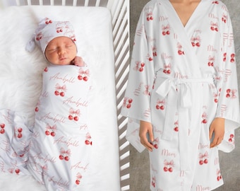May include: A white baby swaddle blanket and matching hat with a cherry and bow pattern, personalized with the name "Anabell". A white robe with a cherry and bow pattern and the word "Mom" is also shown.