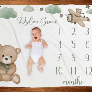 May include: A white baby blanket featuring a green and brown teddy bear, a green aeroplane with a teddy bear pilot, and a green number chart for tracking a baby's growth. The blanket is personalised with the name "Dylan Grant".