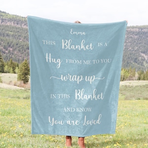 Könnte beinhalten: Eine hellblaue Fleece-Decke mit dem Text "Emma THIS Blanket IS A Hug FROM ME TO YOU wrap up IN THIS Blanket AND KNOW You are Loved" in weiß gedruckt.