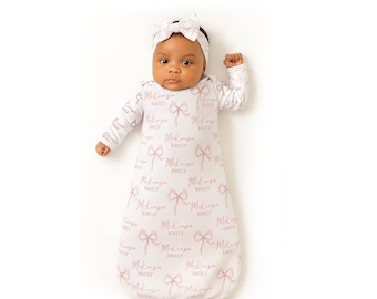 May include: A baby wearing a white sleep sack and matching headband. The sleep sack is patterned with pink bows and the name "McKenzie" and "Emily" in a script font. The baby is lying on a white surface.