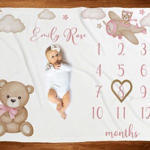 Può includere: Coperta bianca per neonati con orsacchiotto, aereo, nuvole e stelle. La coperta ha il nome "Emily Rose" e i numeri da 1 a 12 con la parola "mesi". Un bambino è sdraiato sulla coperta.