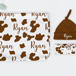 May include: A white baby blanket, hat, and headband set. The blanket and headband feature a brown cow print pattern with the name "Ryan" in a script font. The hat is solid brown with the name "Ryan".