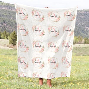 May include: A white blanket with a repeating pattern of pink floral wreaths and the name "Ariana Billy" in a gold circle.