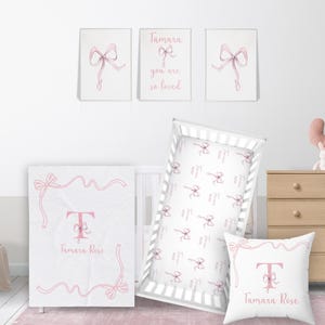 May include: A nursery decor set with a pink and white color scheme. The set includes a crib sheet, a blanket, a pillow, and three wall art prints. The crib sheet and pillow feature a repeating pattern of pink bows. The blanket features a pink bow border and a large pink letter T in the center. The wall art prints feature pink bows and the text "Tamara you are so loved".